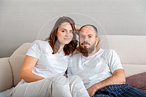 Young beautiful couple in pajamas hug while sitting on couch at cozy home interior. Married man and woman romantic