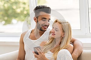 Young Beautiful Couple Sit On Coach Near Window, Using Cell Smart Phone Happy Smile Hispanic Man Woman