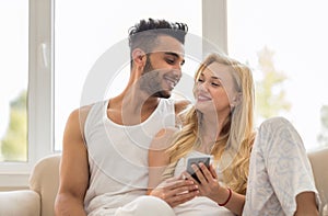 Young Beautiful Couple Sit On Coach Near Window, Using Cell Smart Phone Happy Smile Hispanic Man Woman