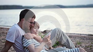 Young beautiful couple sitting on the beach, on the shore of the river and talking, woman using the smartphone.