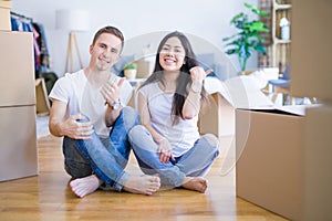 Young beautiful couple sitting on the floor at new home around cardboard boxes smiling with happy face looking and pointing to the