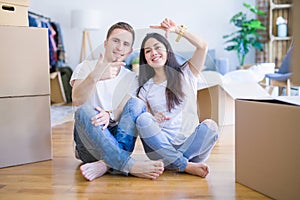 Young beautiful couple sitting on the floor at new home around cardboard boxes smiling making frame with hands and fingers with