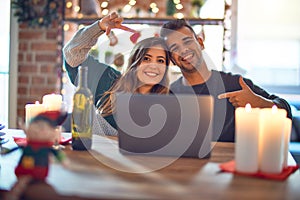 Young beautiful couple sitting using laptop around christmas decoration at home smiling making frame with hands and fingers with