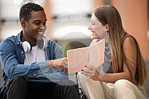 Young beautiful couple smiling and reading together a notebook.Two diverse students sitting outside talking while