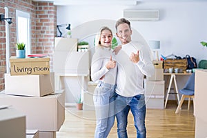 Young beautiful couple standing at new home around cardboard boxes doing happy thumbs up gesture with hand