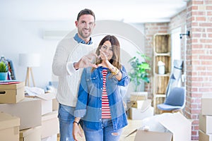 Young beautiful couple standing at new home around cardboard boxes smiling in love doing heart symbol shape with hands