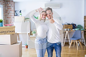 Young beautiful couple standing at new home around cardboard boxes smiling making frame with hands and fingers with happy face