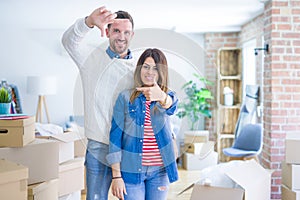 Young beautiful couple standing at new home around cardboard boxes smiling making frame with hands and fingers with happy face