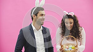 Young beautiful couple standing on a pink background. With hackneyed ears on the head. During this man gives his wife a