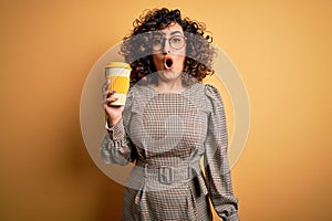 Young beautiful curly arab businesswoman wearing glasses drinking cup of takeaway coffe scared in shock with a surprise face,