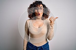 Young beautiful curly arab woman wearing casual t-shirt and glasses over white background Surprised pointing with hand finger to