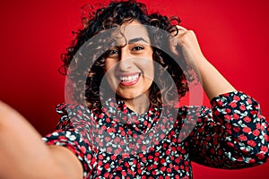 Young beautiful curly arab woman wearing dress making selfie by camera over red background very happy pointing with hand and