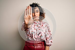 Young beautiful curly arab woman wearing floral t-shirt standing over isolated white background doing stop sing with palm of the