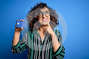 Young beautiful curly arab woman wearing glasses holding credit card money for payment serious face thinking about question, very