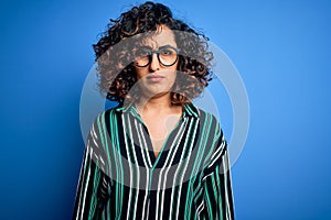 Young beautiful curly arab woman wearing striped shirt and glasses over blue background skeptic and nervous, frowning upset