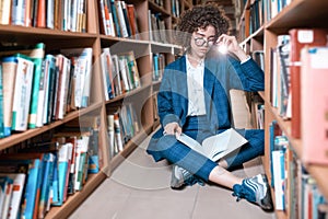 Young beautiful curly girl in glasses and blue suit sitting with books in the library.