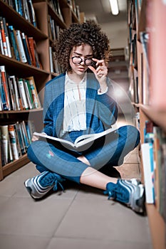 Young beautiful curly girl in glasses and blue suit sitting with books in the library.