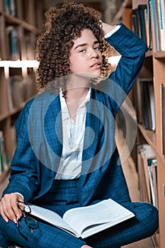 Young beautiful curly girl in glasses and blue suit sitting with books in the library.