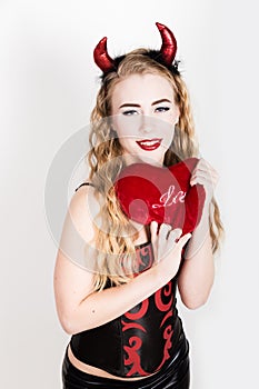 Young and beautiful curly girl with red horns looks like pretty Devil, holding a heart pillow