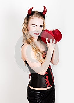 Young and beautiful curly girl with red horns looks like pretty Devil, holding a heart pillow