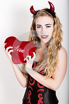 Young and beautiful curly girl with red horns looks like pretty Devil, holding a heart pillow