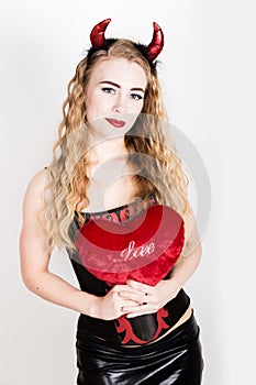 Young and beautiful curly girl with red horns looks like pretty Devil, holding a heart pillow