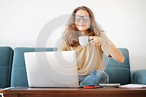Young beautiful curly woman with glasses enjoying morning coffee in front of a laptop screen at home on a blue sofa