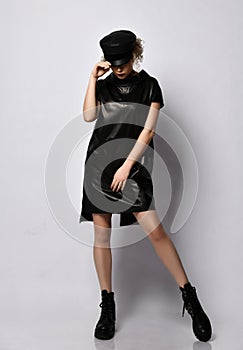 Young beautiful curly woman in stylish black leather dress, cap and brutal boots stands looking down