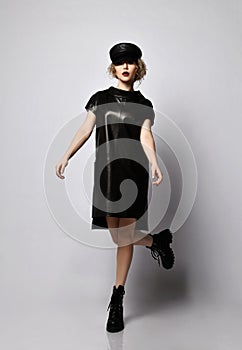 Young beautiful curly woman in stylish black leather dress, hat and massive boots walking over grey wall background