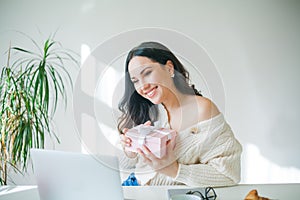 Young beautiful dark-haired woman of 34 years old in knitted sweater is talking on video link and holding pink gift box in her