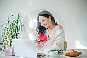 Young beautiful dark-haired woman of 34 years old in knitted sweater is talking on videophone and holding plush heart in her hands