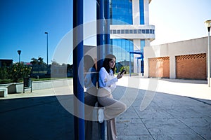 Young, beautiful, dark-haired woman is consulting her mobile phone at the entrance door of the building where she works as an