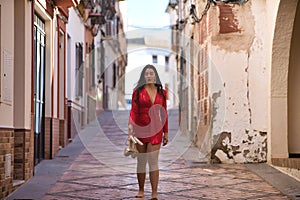 Young, beautiful and dark-haired woman in an elegant red dress and heels in her hand, walking barefoot, at dawn along a lonely