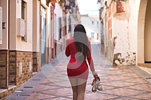 Young, beautiful, dark-haired woman, with her back turned, wearing an elegant red dress and heels in her hand, walking barefoot,