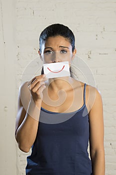 Young beautiful depressed woman hiding her sorrow and sadness behind a paper with a drawn smile