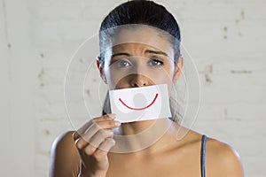 Young beautiful depressed woman hiding her sorrow and sadness behind a paper with a drawn smile photo