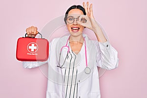 Young beautiful doctor woman with blue eyes wearing stethoscope holding first aid kit box with happy face smiling doing ok sign