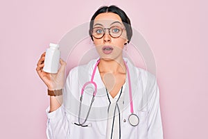 Young beautiful doctor woman with blue eyes wearing stethoscope holding medicine pills scared in shock with a surprise face,