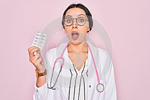 Young beautiful doctor woman with blue eyes wearing stethoscope holding medicine pills scared in shock with a surprise face,
