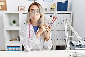 Young beautiful doctor woman holding diapason instrument puffing cheeks with funny face