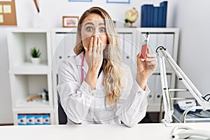 Young beautiful doctor woman holding electronic cigarette at the clinic covering mouth with hand, shocked and afraid for mistake