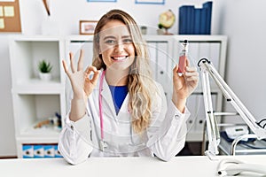 Young beautiful doctor woman holding electronic cigarette at the clinic doing ok sign with fingers, smiling friendly gesturing