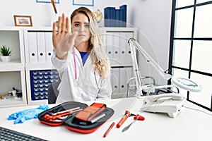 Young beautiful doctor woman with reflex hammer and medical instruments doing stop sing with palm of the hand