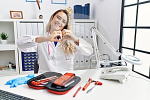 Young beautiful doctor woman with reflex hammer and medical instruments smiling in love doing heart symbol shape with hands