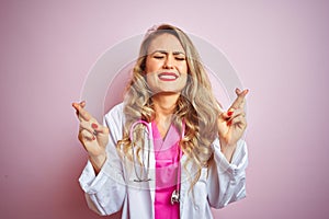 Young beautiful doctor woman using stethoscope over pink isolated background gesturing finger crossed smiling with hope and eyes