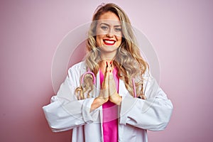 Young beautiful doctor woman using stethoscope over pink isolated background praying with hands together asking for forgiveness