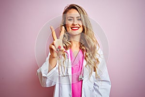 Young beautiful doctor woman using stethoscope over pink isolated background showing and pointing up with fingers number two while