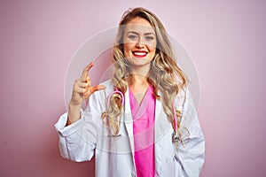 Young beautiful doctor woman using stethoscope over pink isolated background smiling and confident gesturing with hand doing small
