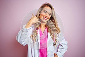 Young beautiful doctor woman using stethoscope over pink isolated background smiling doing phone gesture with hand and fingers