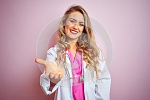 Young beautiful doctor woman using stethoscope over pink isolated background smiling friendly offering handshake as greeting and